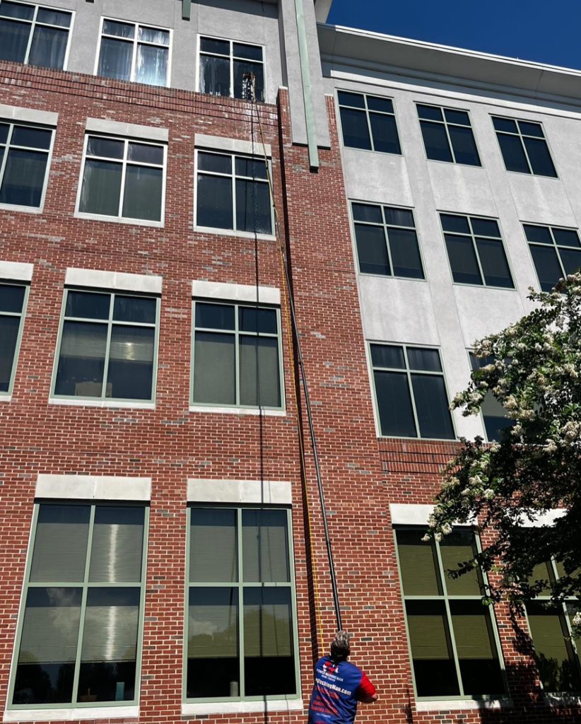 A person in a red and blue shirt stands watching a fire escape ladder descend from a multistory, red brick and gray building on a sunny day.