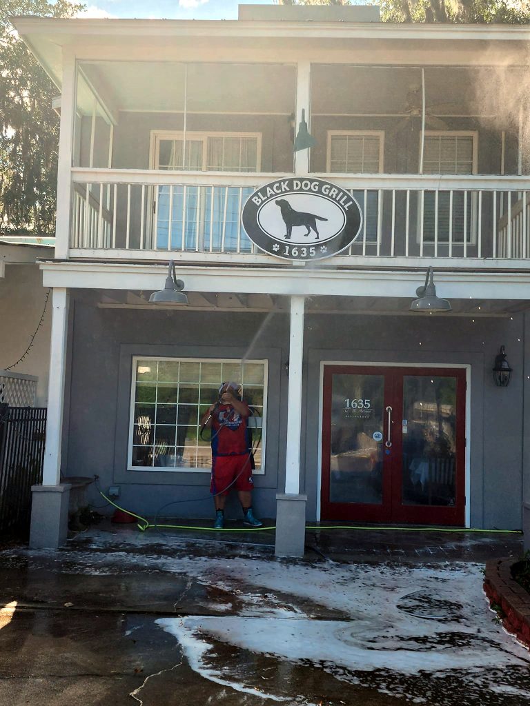 A person in a red shirt and shorts is washing the sidewalk in front of the "Black Dog Grill" restaurant with a hose.