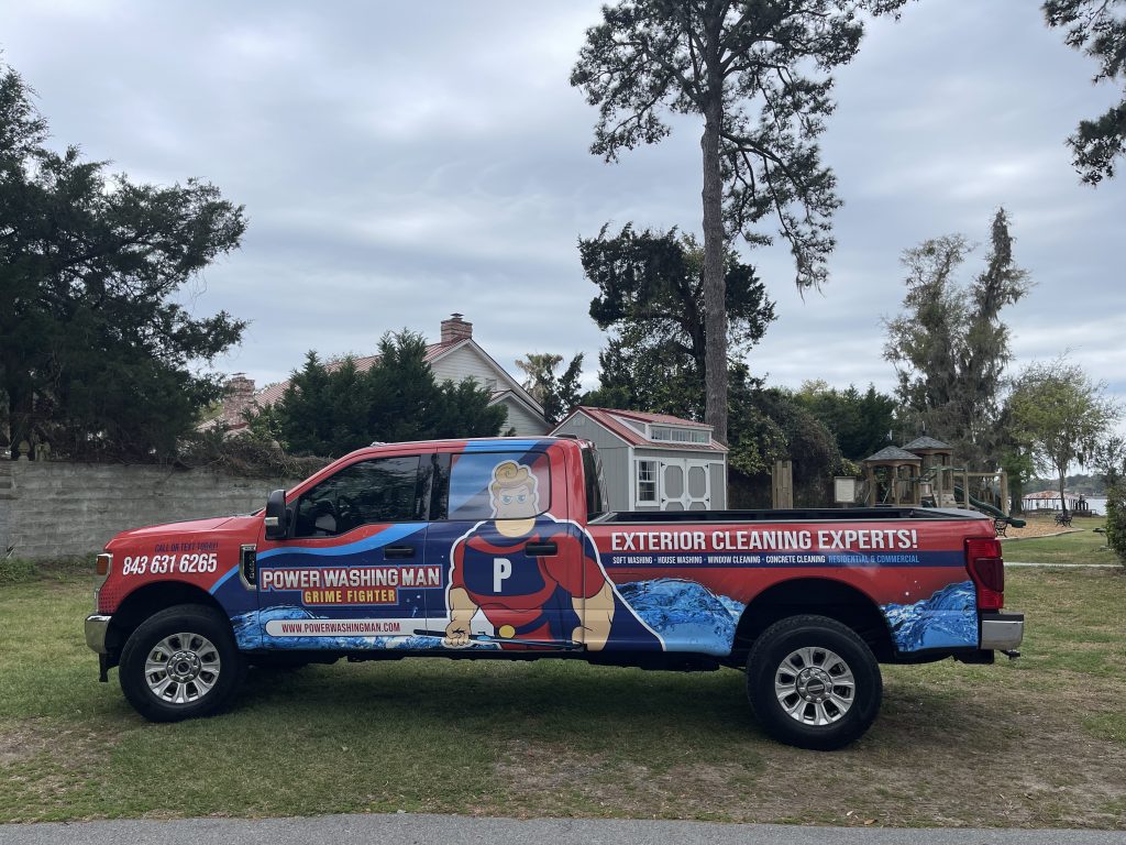 A red pickup truck with advertising graphics for a power washing service, featuring a superhero, parked in a grassy area near trees.