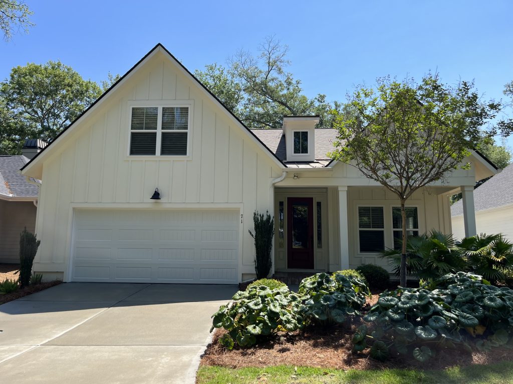 A suburban single-story house with a gabled roof, attached garage, a red door, and landscaped with greenery under a clear blue sky.