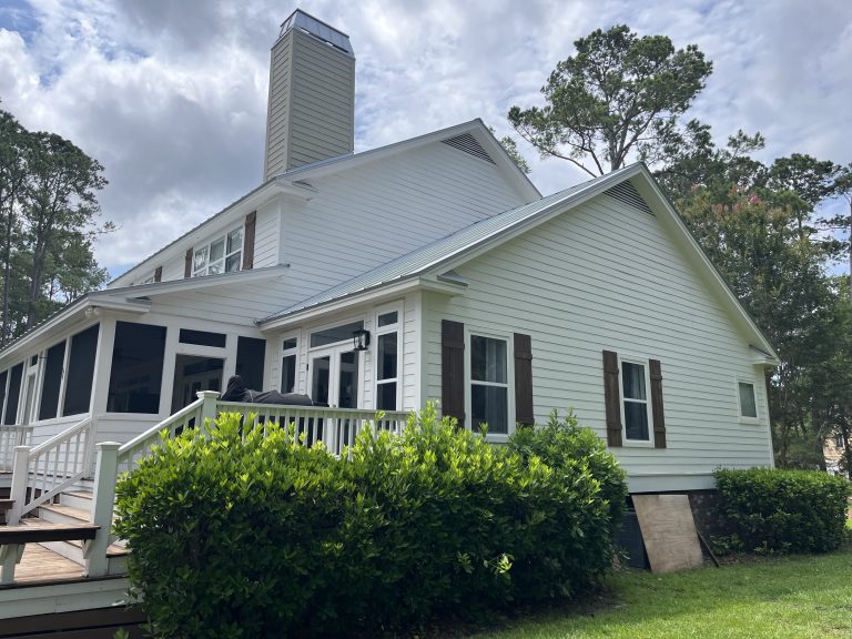A two-story house with pale siding, a large chimney, screened porch, and wooden stairs surrounded by greenery under a partly cloudy sky.