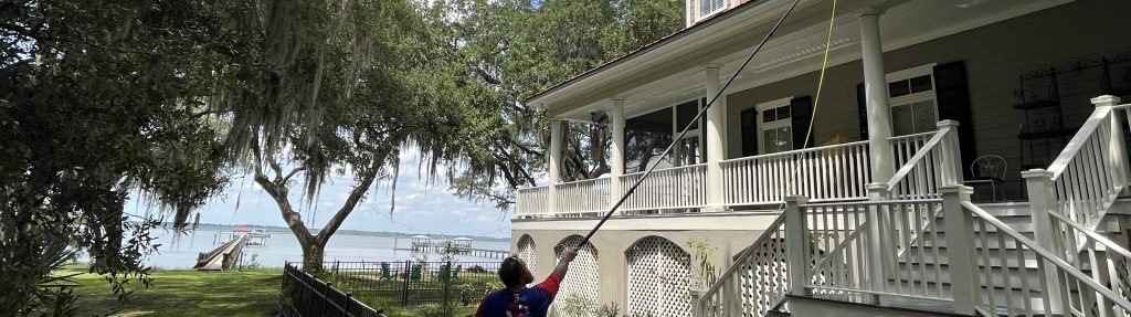A person is holding a fishing rod near a large white house with a balcony, surrounded by trees, overlooking a tranquil water body with a pier.