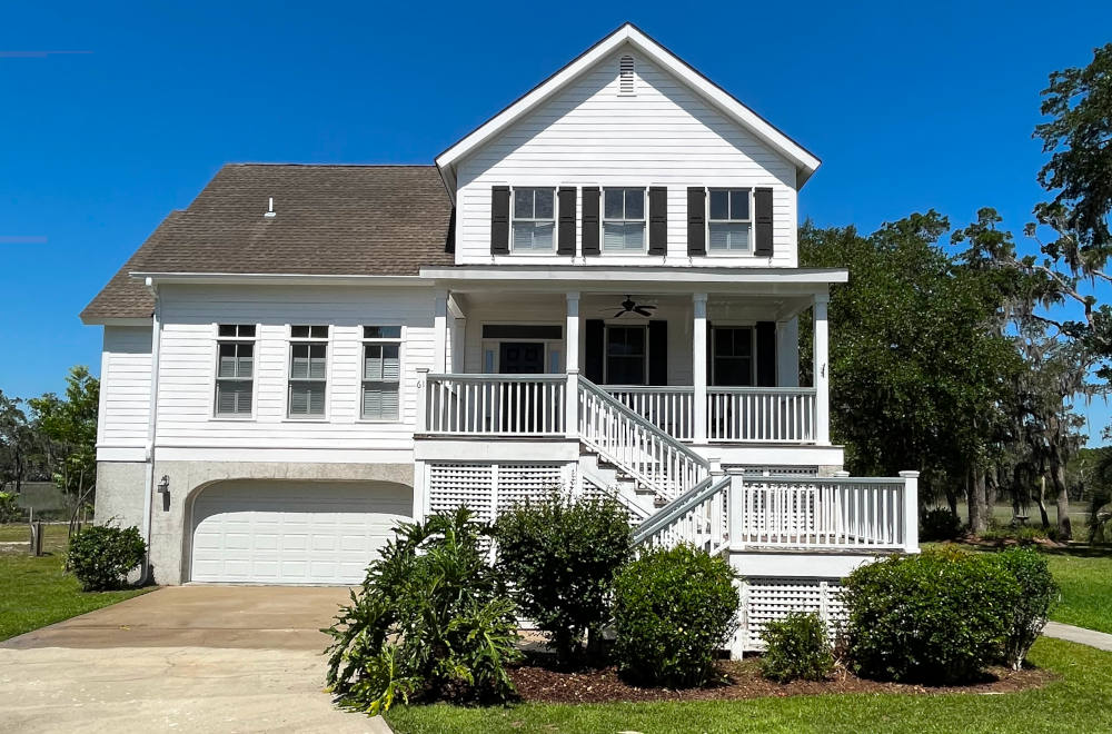 A two-story white house with a balcony, garage, and staircase leading to a raised entrance. Landscaped with bushes, under a clear blue sky.