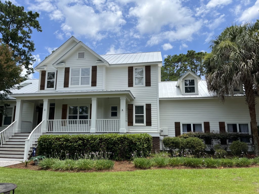 A two-story white house with a covered front porch, gabled roof, palm tree, green lawn, and a clear blue sky with fluffy clouds above.